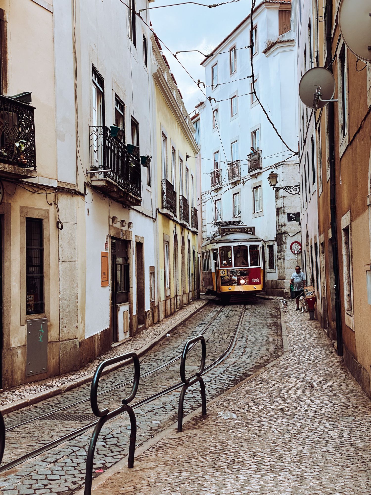 A vintage red tram navigates a narrow cobblestone street in Lisbon, Portugal, flanked by traditional white buildings.