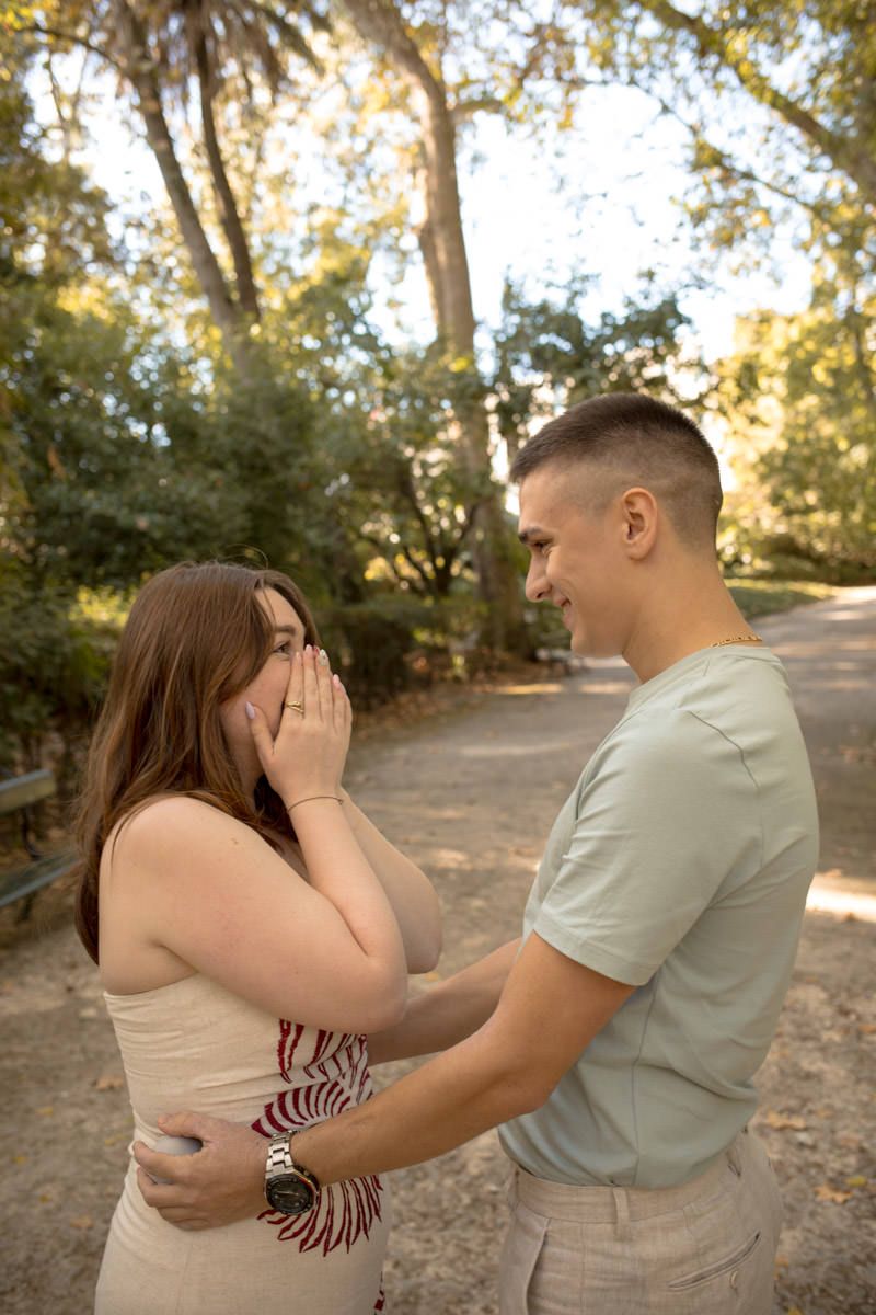 A couple shares a joyful surprise moment in a sunlit park, woman covering her mouth in excitement.