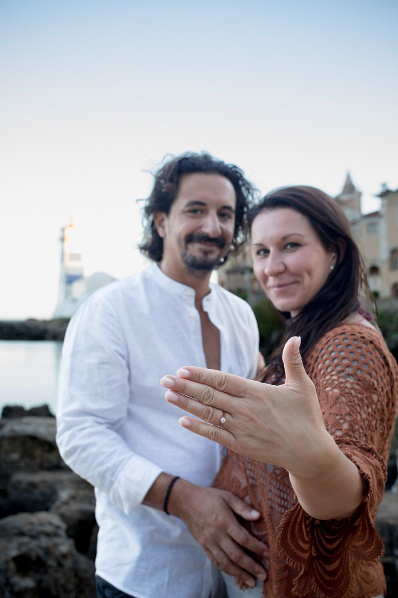Happy couple showing off engagement ring outdoors near waterfront at dusk, woman extending hand toward camera.
