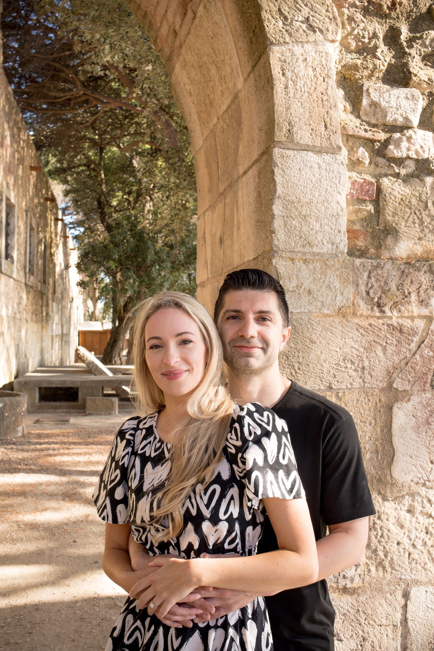Couple smiling together under stone arches, woman in heart-print dress, man in black shirt, outdoor historic setting.