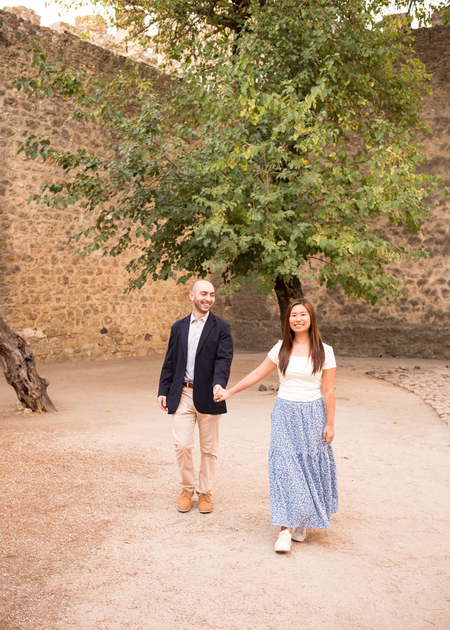 A couple holds hands and walks together outdoors near a stone wall with a large green tree behind them.