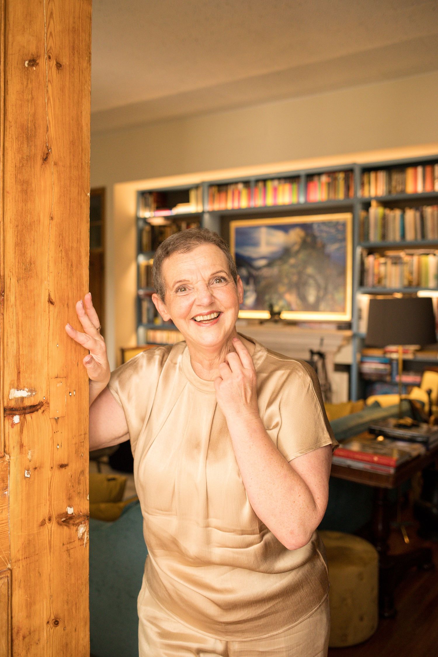 Smiling senior woman in gold blouse standing by wooden door in cozy home library.