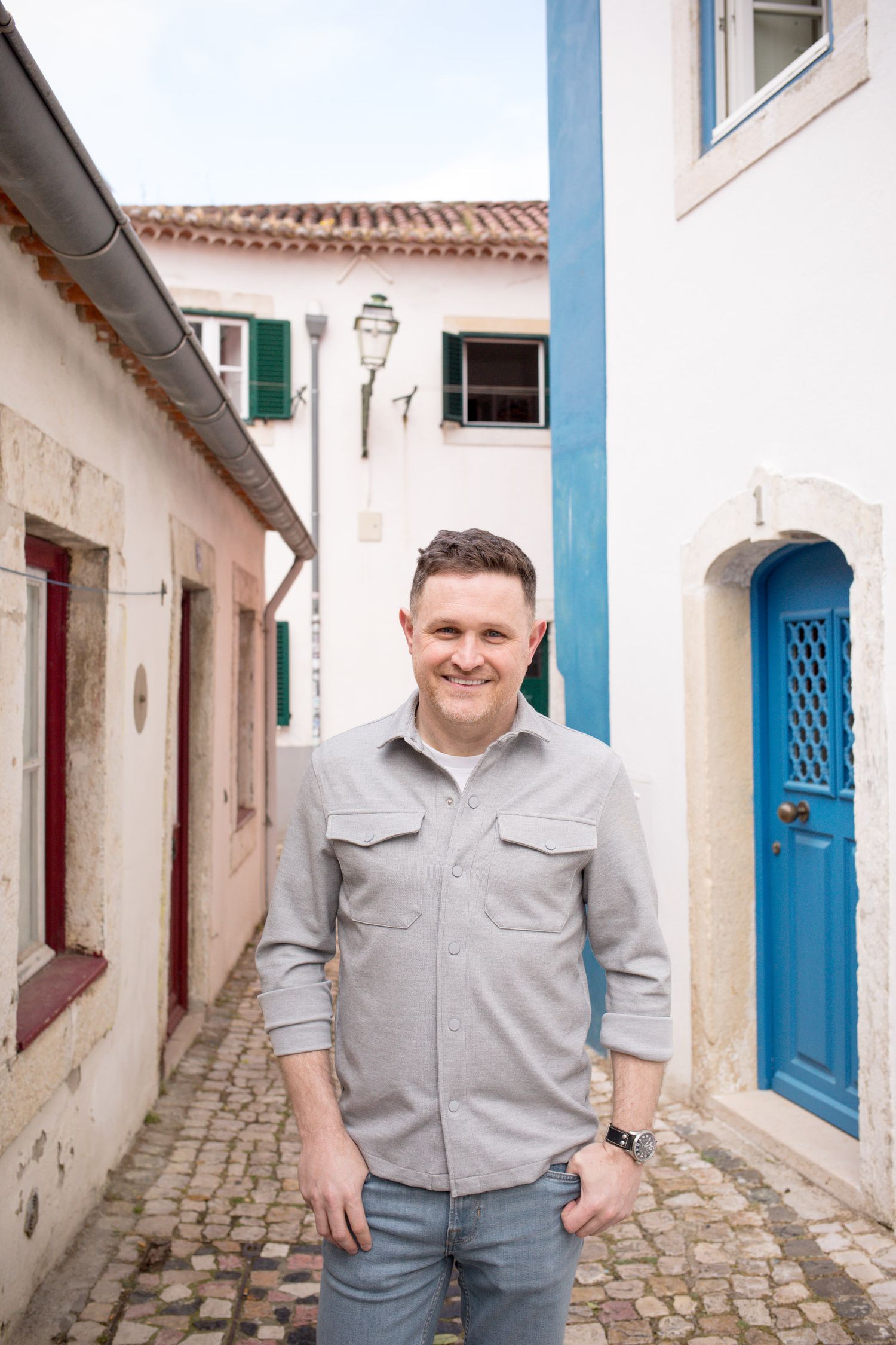 Smiling man in grey shirt standing on cobblestone alley between white European buildings.