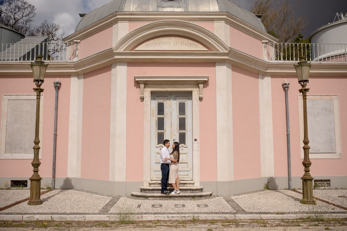 Couple embracing in front of a pink neoclassical building with white door and ornate columns.