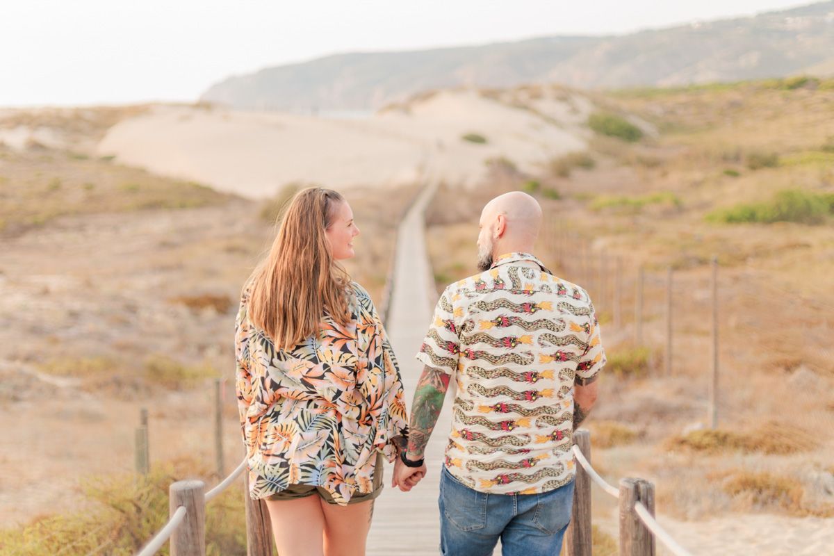 Couple holding hands walking on a wooden boardwalk through sandy dunes, viewed from behind.