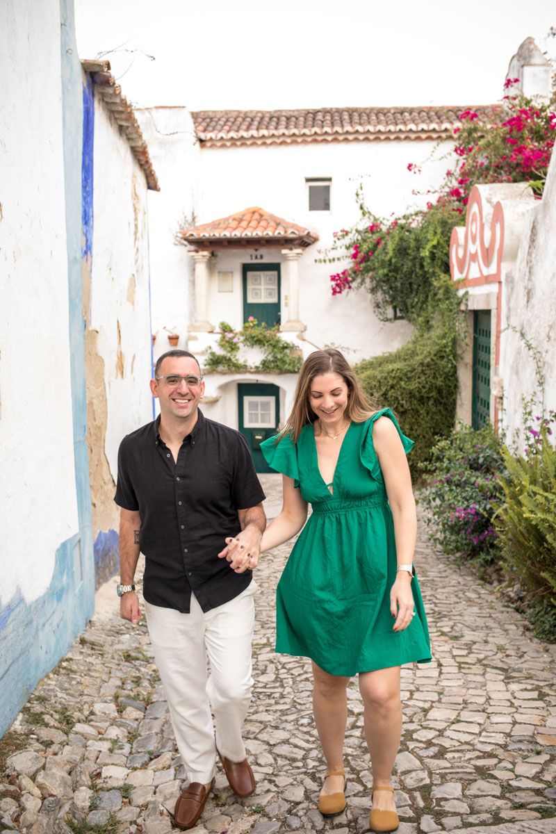 Couple holding hands walking down a charming cobblestone alley with white buildings and colorful flowers.