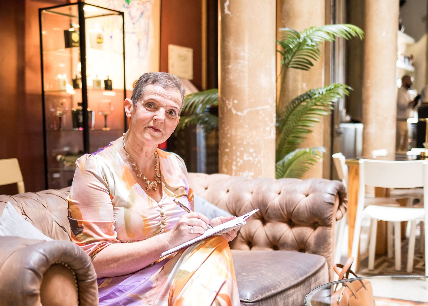 Woman with short hair and glasses sits on a tufted brown leather sofa writing in a notebook in an elegant lounge with potted ferns.