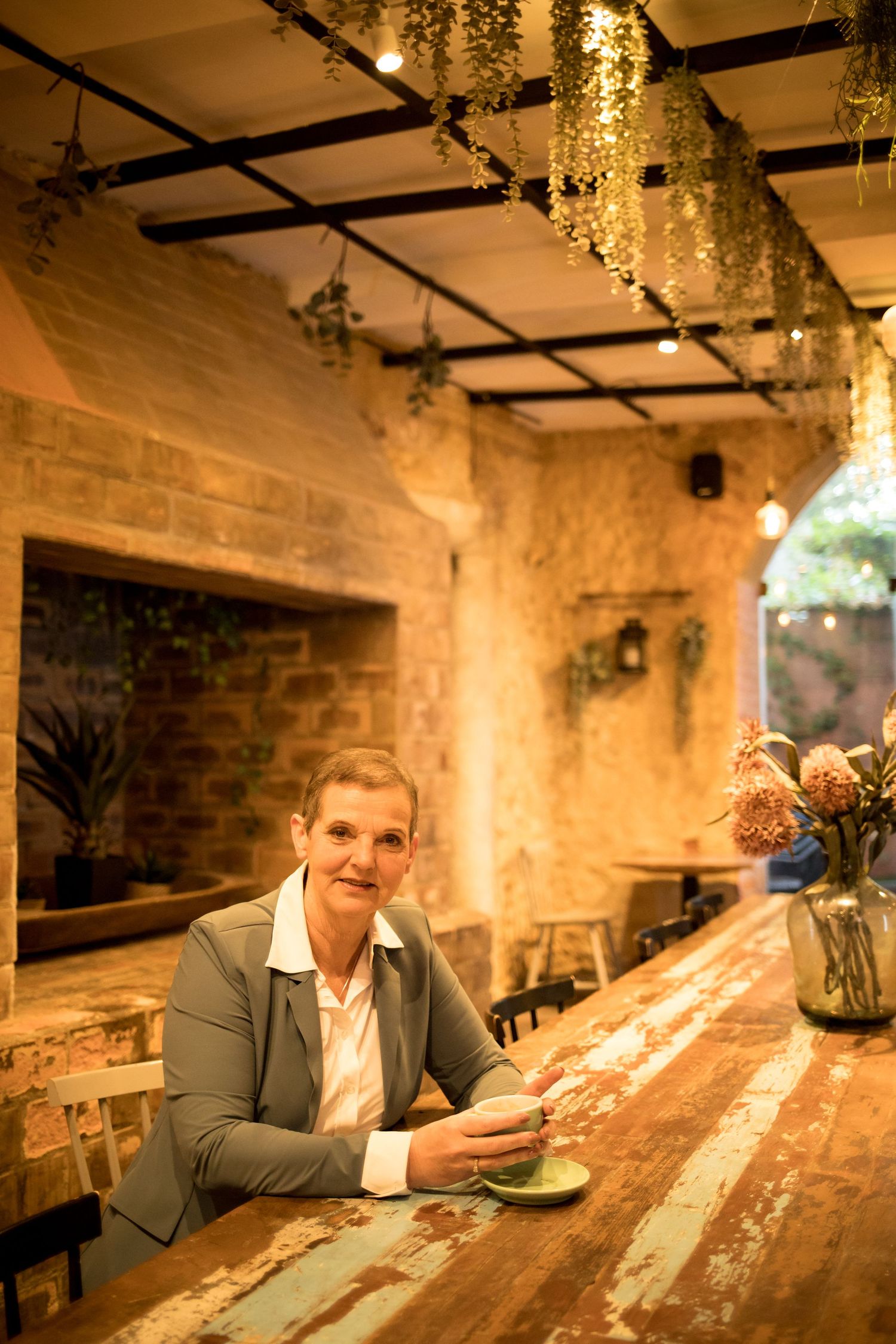 Woman in a gray blazer sits at a rustic wooden table holding a green teacup in a cozy brick cafe with hanging plants.
