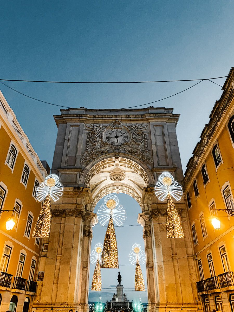 Rua Augusta Arch in Lisbon adorned with Christmas lights and trees at dusk, with golden buildings flanking the triumphal arch.
