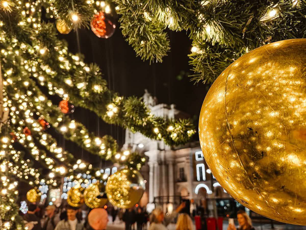 Glowing golden Christmas ornaments and twinkling lights adorn a festive outdoor tree at a holiday market.