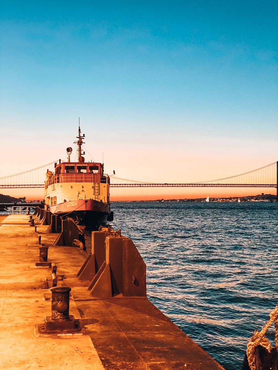 A tugboat docked at a sunlit waterfront pier with a suspension bridge and calm blue waters at golden hour.