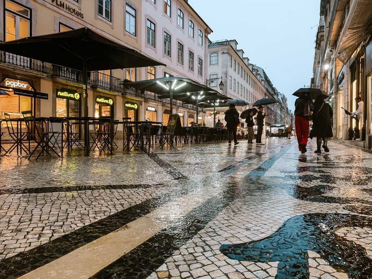 Rainy evening on a cobblestone street in Lisbon, Portugal, with wet reflections and pedestrians carrying umbrellas near outdoor cafes.