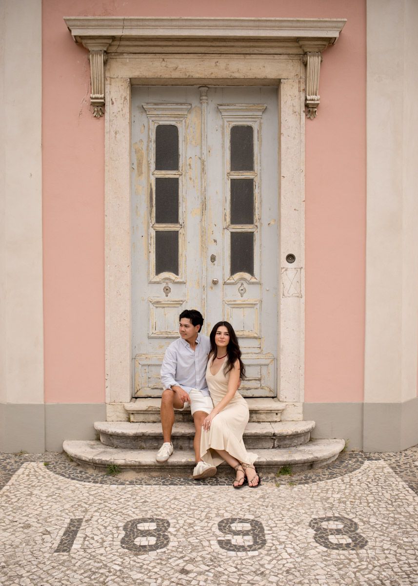 Couple sitting on steps of a vintage pink building with ornate door and cobblestone year 1898 in Lisbon.