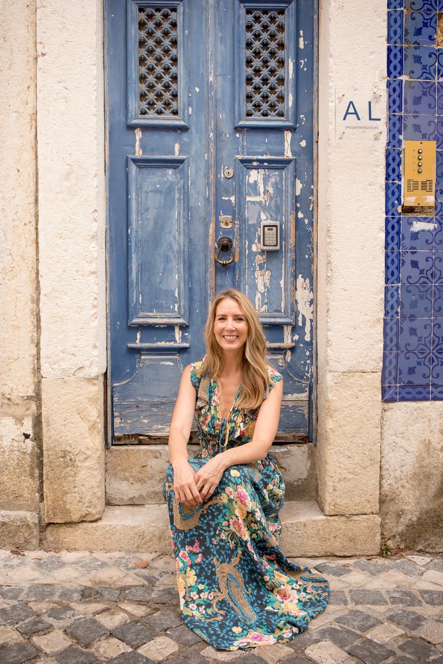 Smiling woman in floral dress sitting before weathered blue door with decorative tiles.