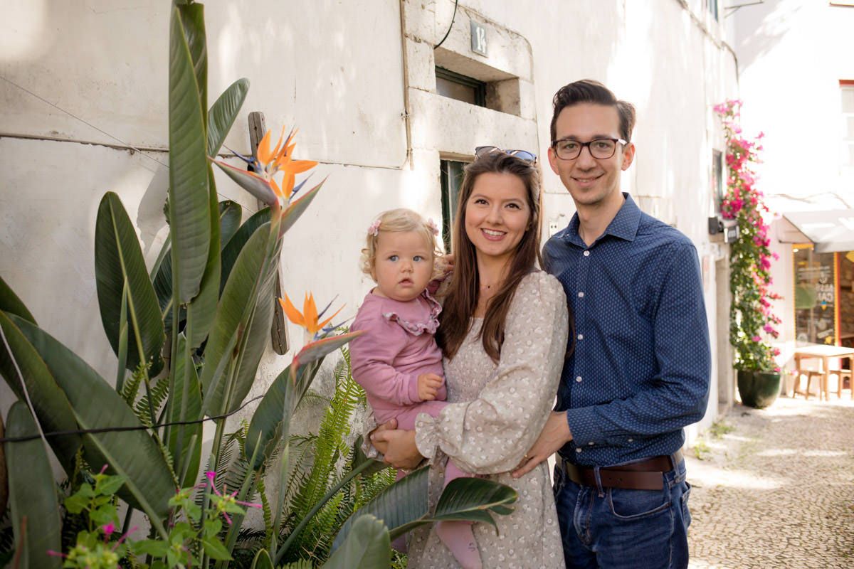 Young family with baby poses beside tropical bird of paradise plants against a white Mediterranean building wall.