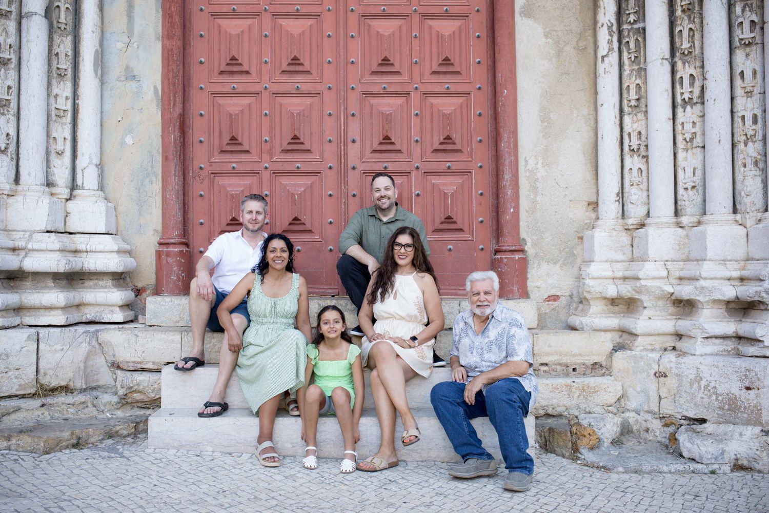 A multigenerational family sits together on stone steps in front of a tall red historic doorway during a Lisbon family photoshoot.