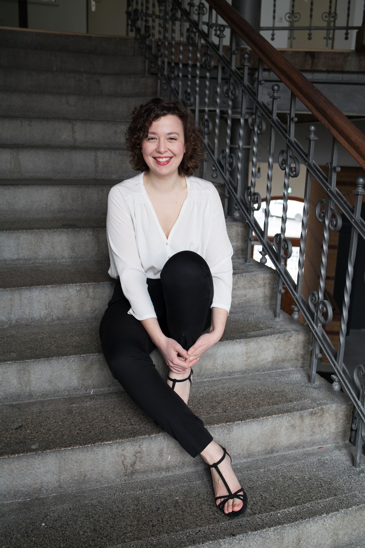 Smiling woman with short curly hair in a white blouse and black pants sits on worn stone stairs beside an ornate wrought iron railing.