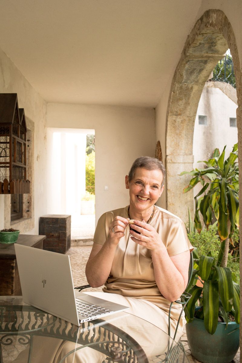 Smiling senior woman sitting at glass table with laptop in elegant arched Mediterranean-style patio surrounded by plants.