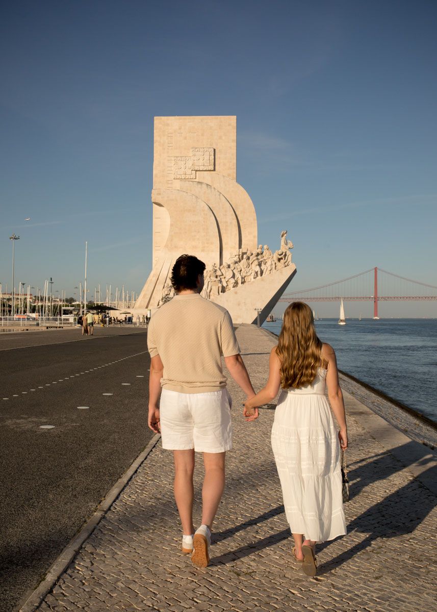Couple holding hands walking toward the Monument to the Discoveries in Lisbon, Portugal on a sunny day.