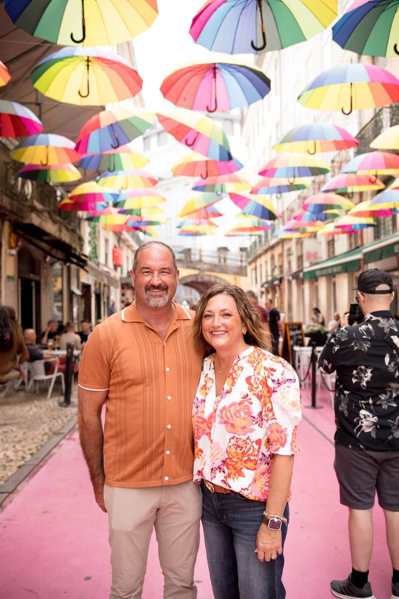 Couple smiling under colorful rainbow umbrellas on a vibrant pink street in Lisbon, Portugal.