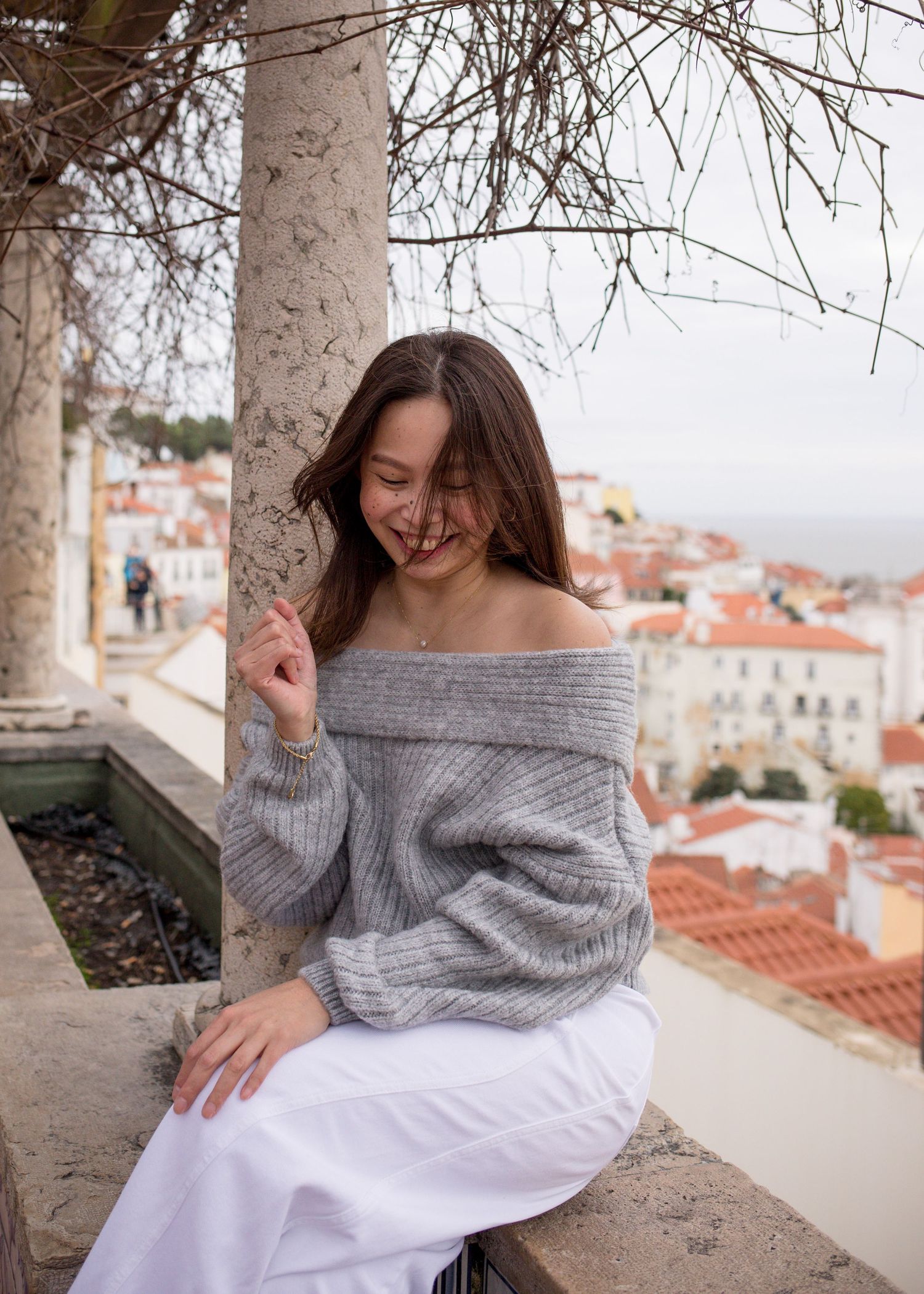 Young woman in gray off-shoulder sweater smiling outdoors with Lisbon cityscape in background.