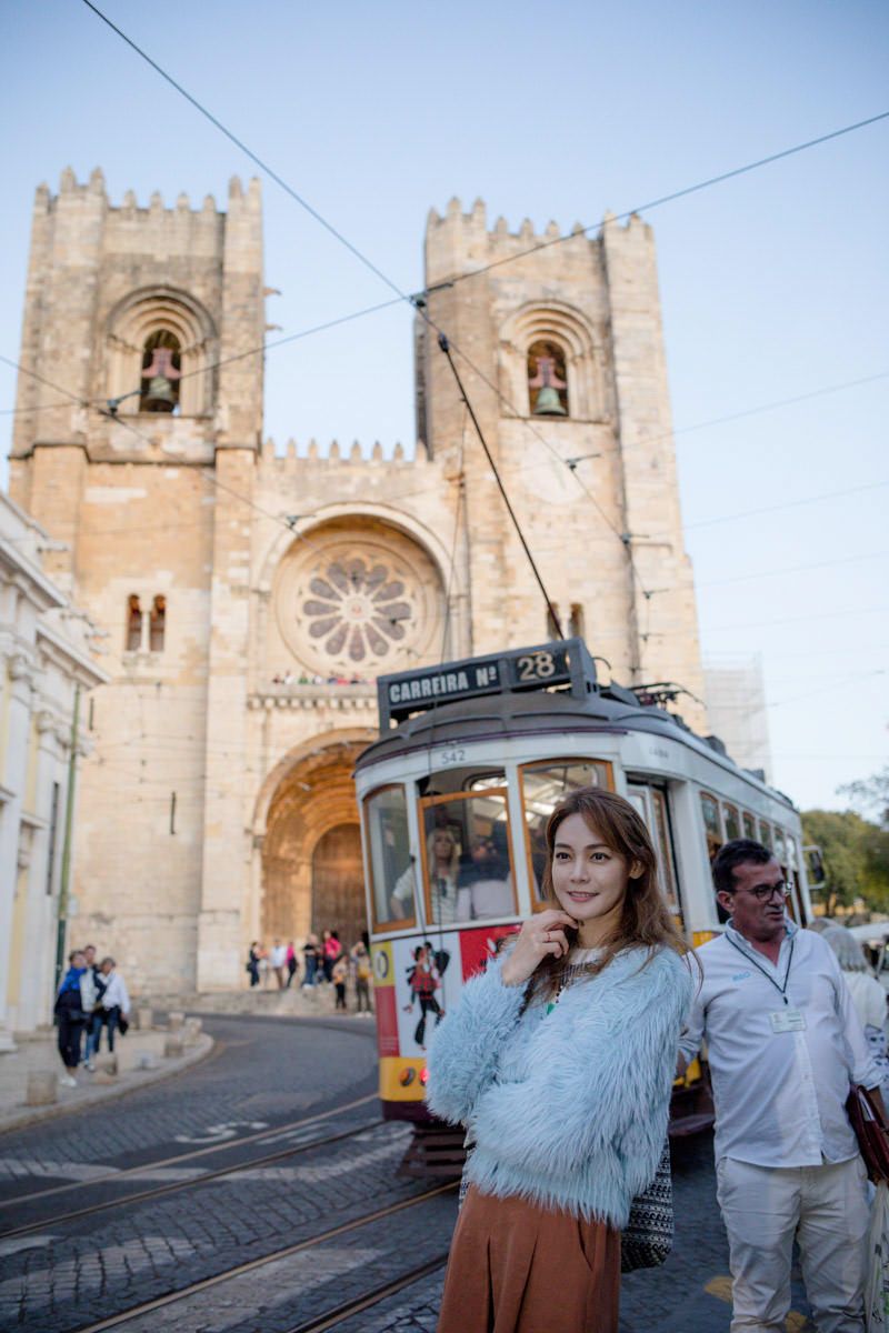 Young woman in fuzzy sweater poses before Lisbon Cathedral with iconic yellow tram 28 on cobblestone street.