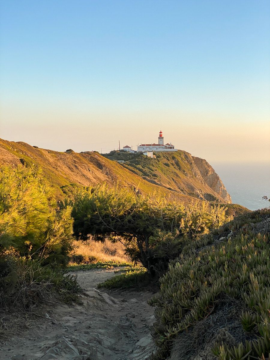 A sandy trail winds through coastal shrubs toward a red and white lighthouse perched on a sunlit cliff above the ocean.