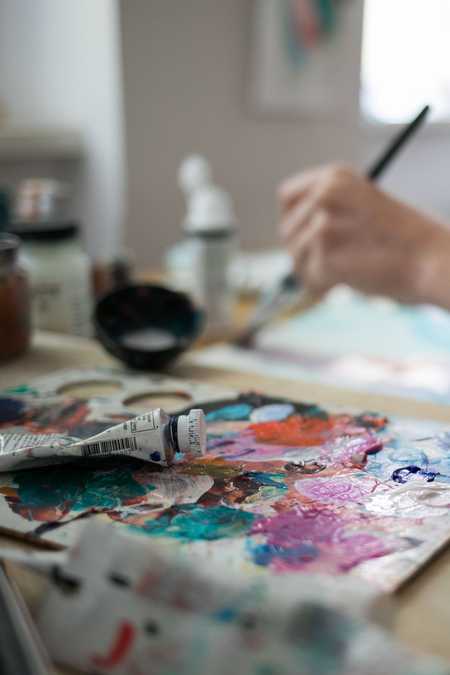 Artist painting with a brush over a colorful abstract watercolor palette, with a paint tube and art supplies on a sunlit studio table.