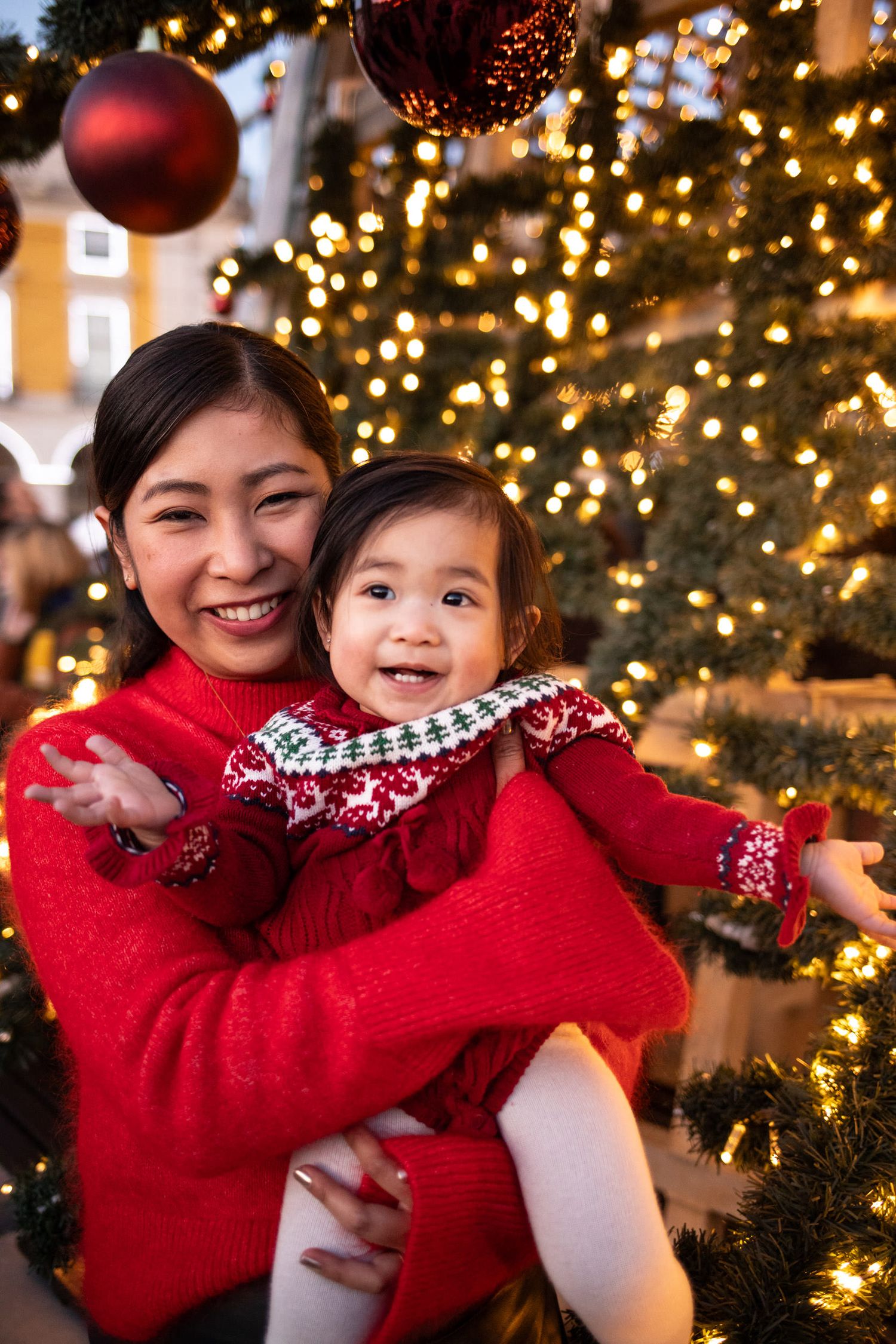A smiling Asian mother in a red sweater holds her toddler daughter near a decorated Christmas tree with warm lights.