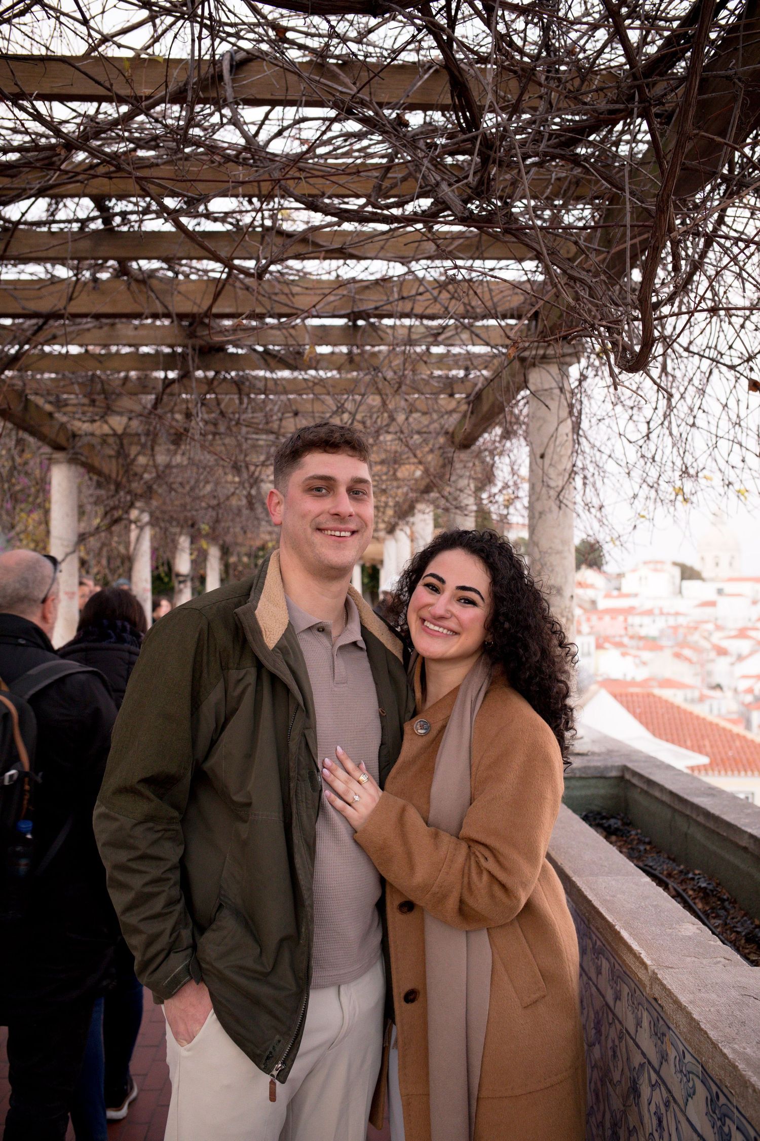 A smiling couple poses under a vine-covered pergola, woman in tan coat resting hand on man in green jacket.