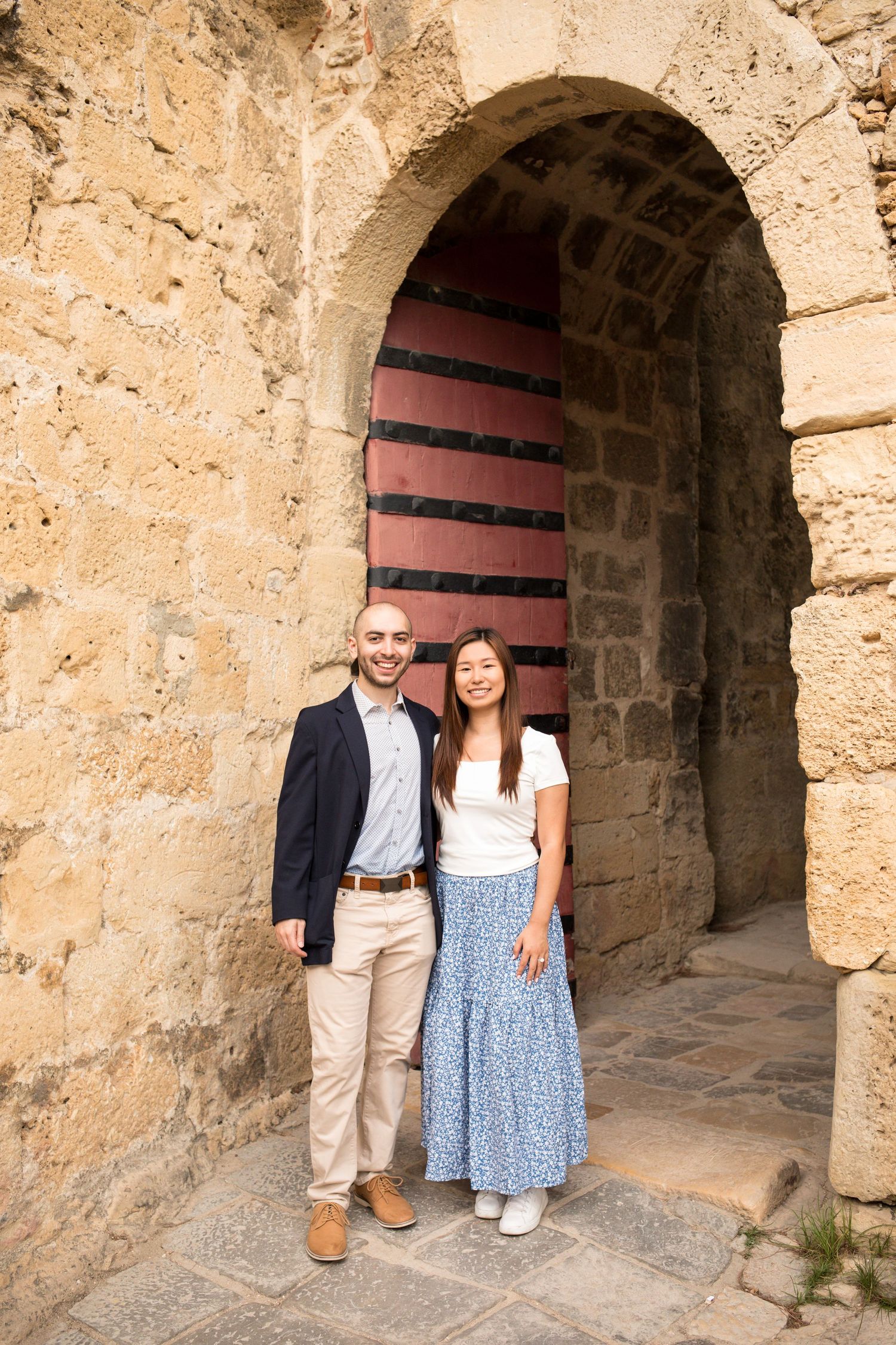 Couple smiling together under a stone archway with a red wooden door at a historic landmark.