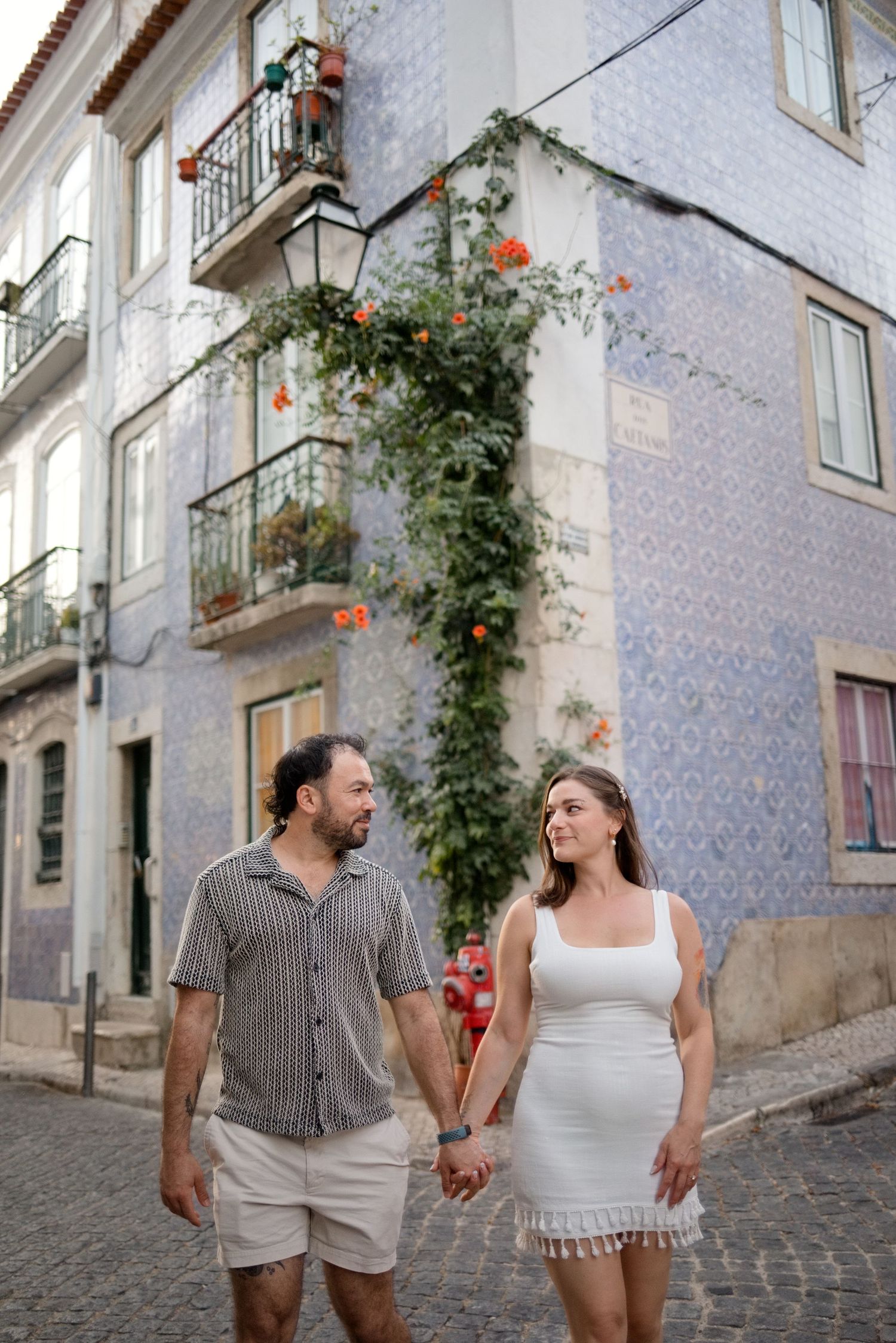 Couple holding hands walking on cobblestone street in Lisbon, Portugal, with blue tiled buildings and orange flowers.
