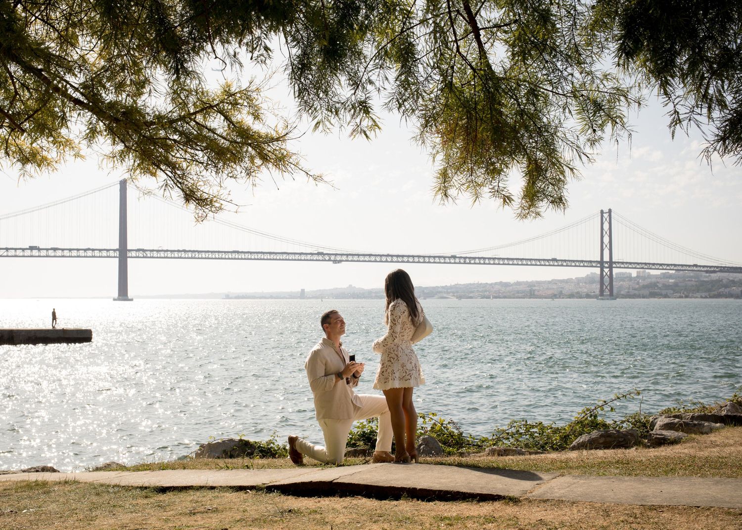Couple sharing a marriage proposal by the Tagus River with the 25 de Abril Bridge in Lisbon, Portugal.