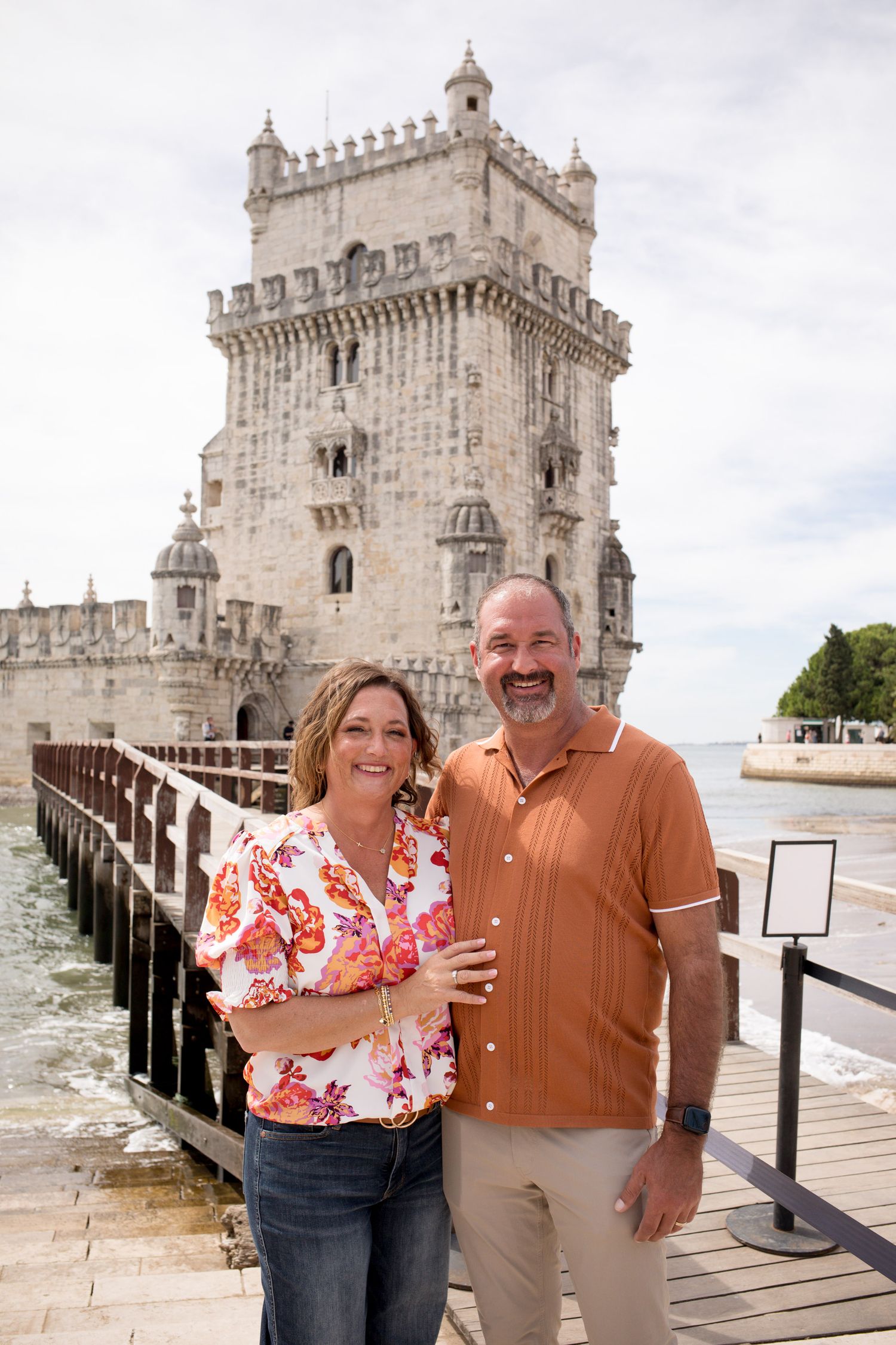 Smiling couple posing in front of the historic Torre de Belem tower along the Lisbon waterfront.