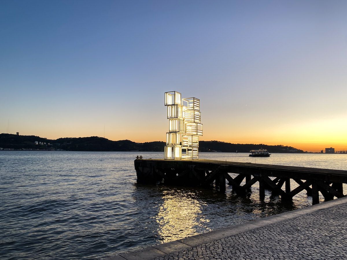Illuminated modern sculpture tower on a pier at sunset over calm water with a colorful sky in the background.