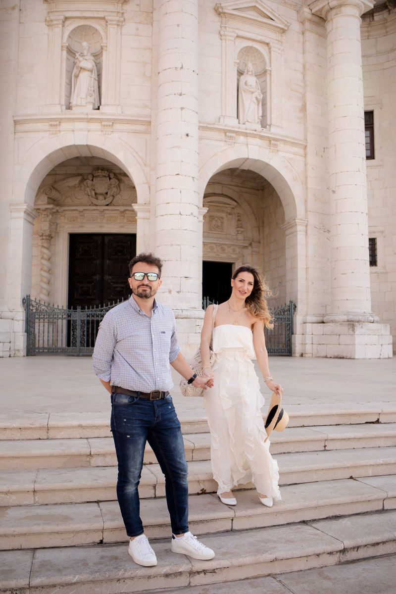 Couple holding hands on church steps, woman in white dress, man in jeans and sunglasses, classic European architecture.