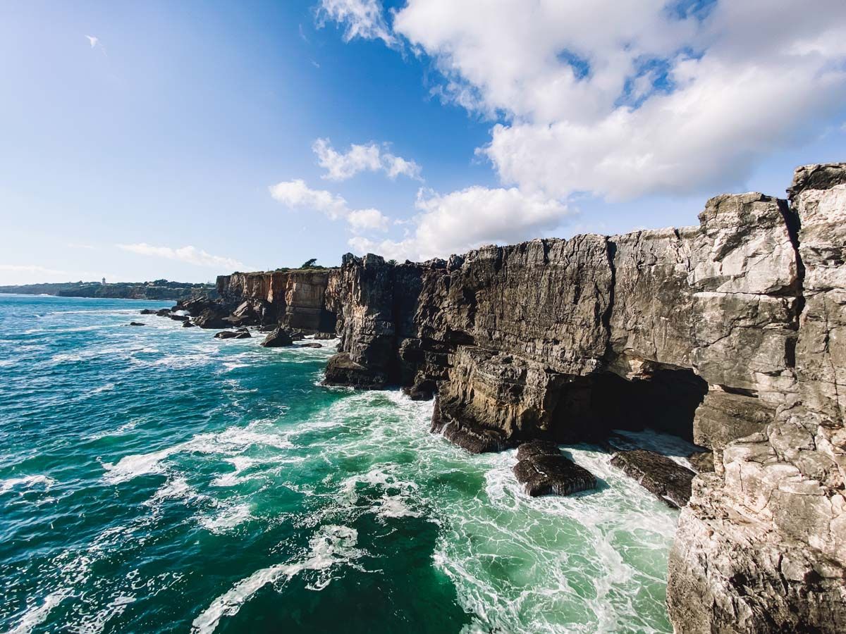Rocky coastal cliffs with turquoise ocean waves crashing below under a partly cloudy blue sky.