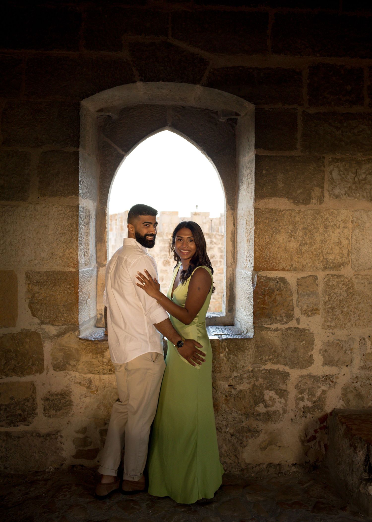 Couple embracing in front of a Gothic arched window in a historic stone castle, woman in green dress.