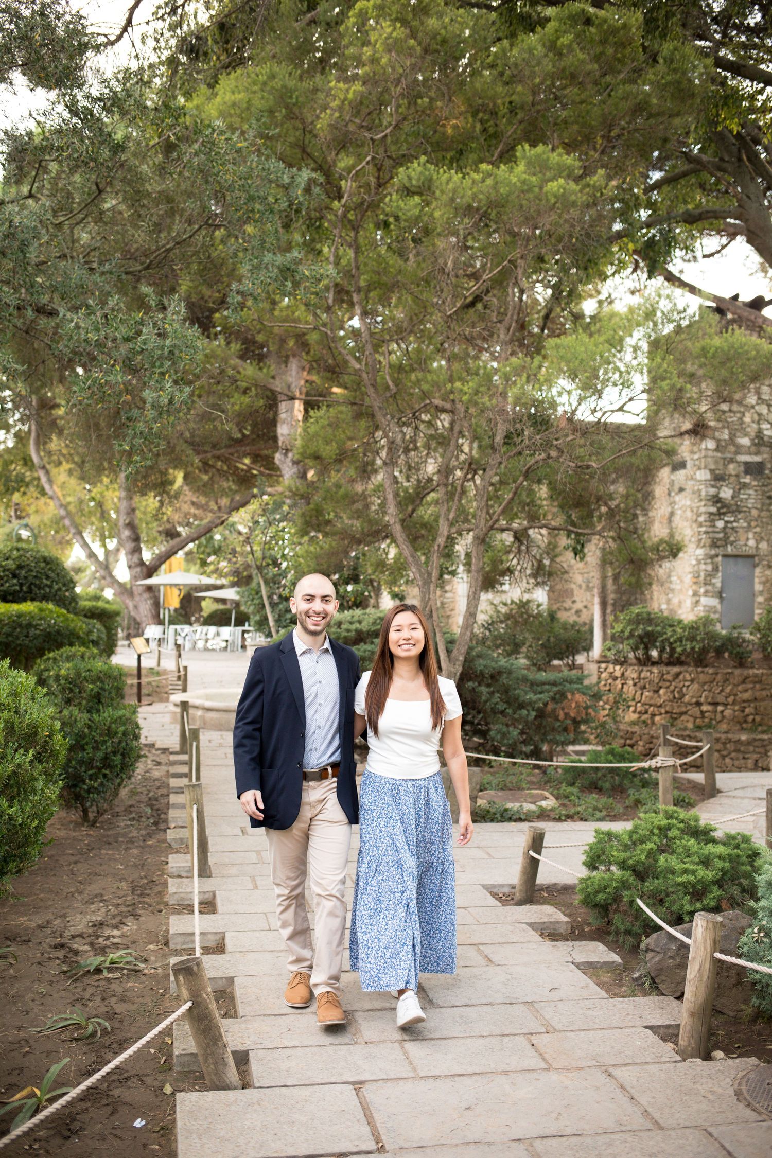Couple walking hand-in-hand on a stone path surrounded by lush green trees and manicured shrubs.