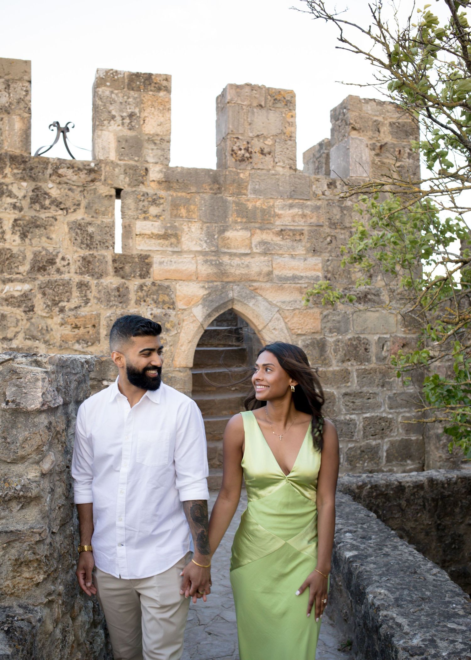 Couple holding hands smiling at each other by ancient stone castle walls, man in white shirt, woman in green dress.