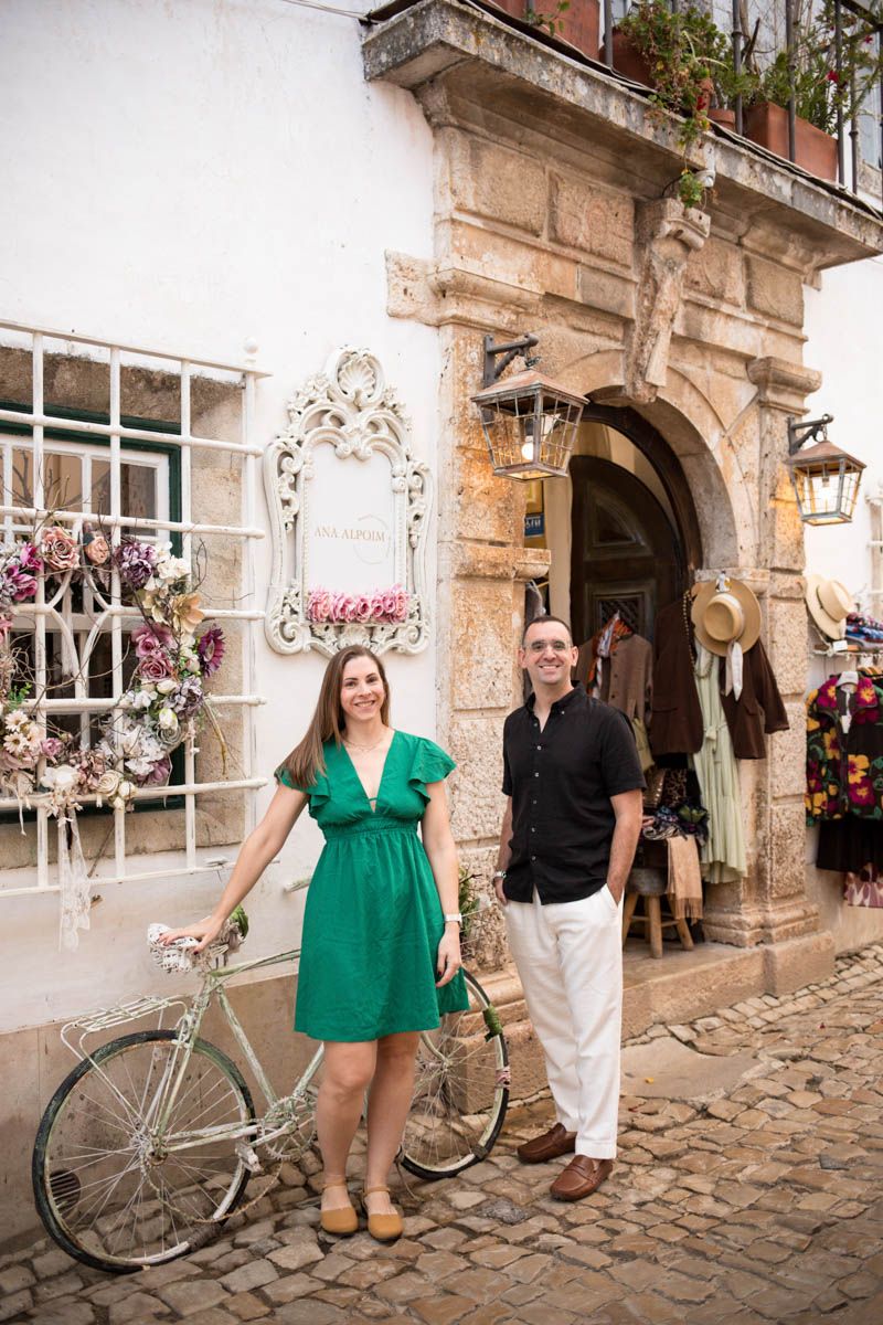 Couple smiling in front of a charming European shop with a white bicycle and cobblestone street.