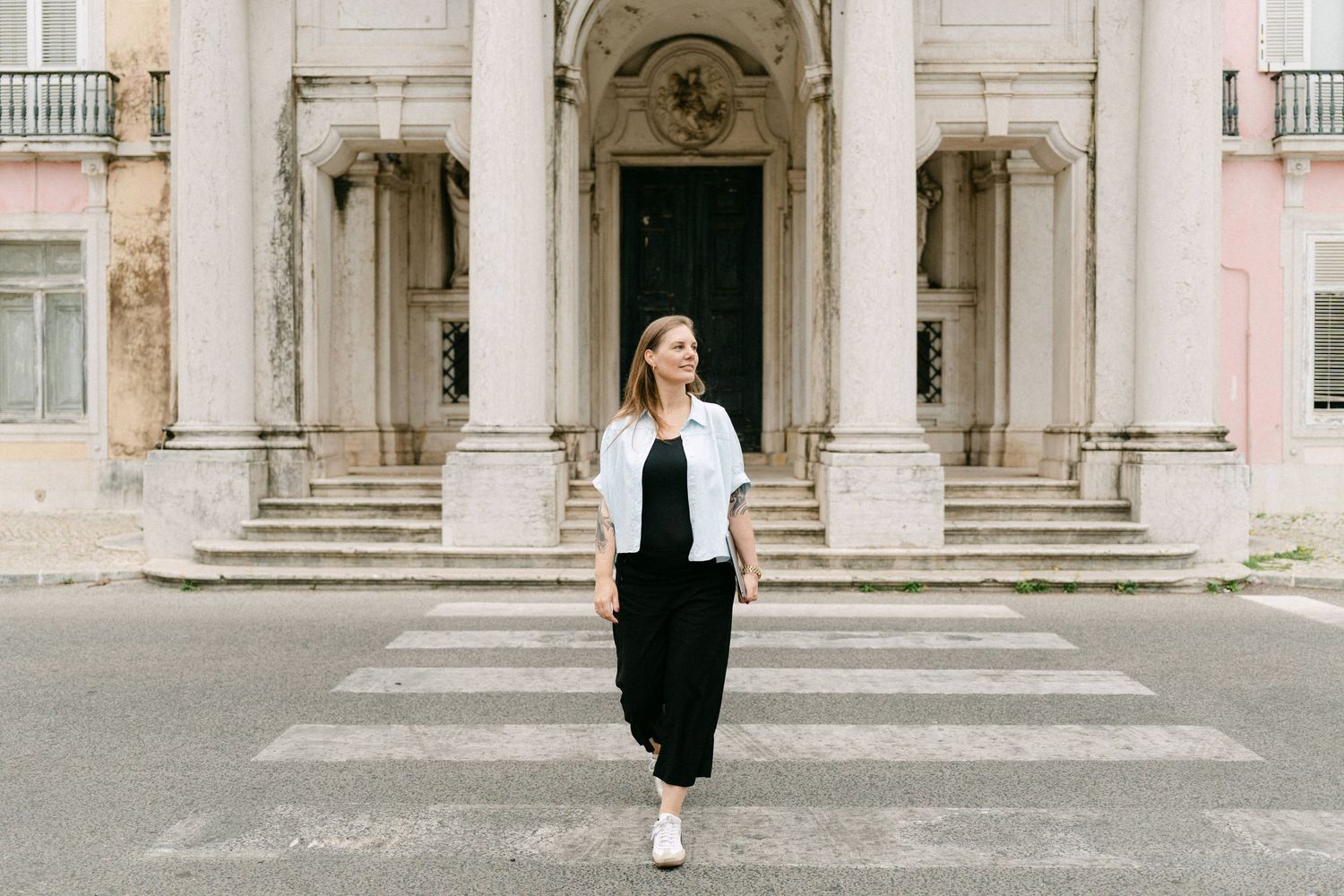 Woman in black outfit and white blazer walking on a crosswalk in front of a grand historic building.