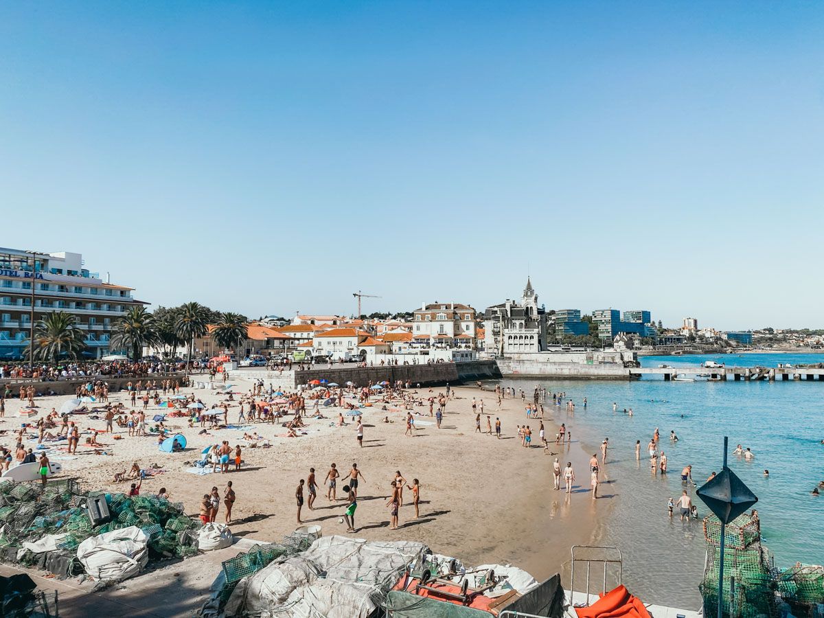 Crowded sandy beach with swimmers and sunbathers along a scenic coastal town waterfront on a clear sunny day.