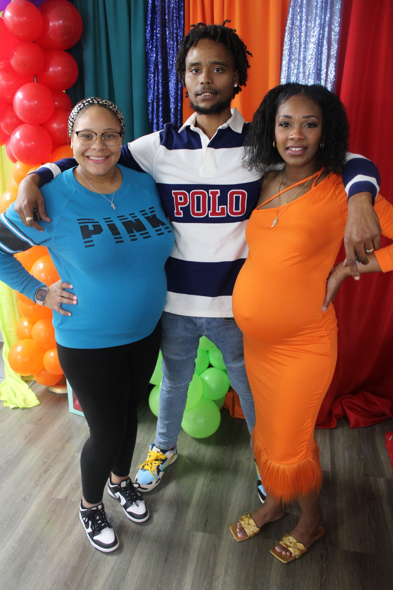 Three people pose at colorful balloon-decorated baby shower, woman in orange dress showing baby bump.