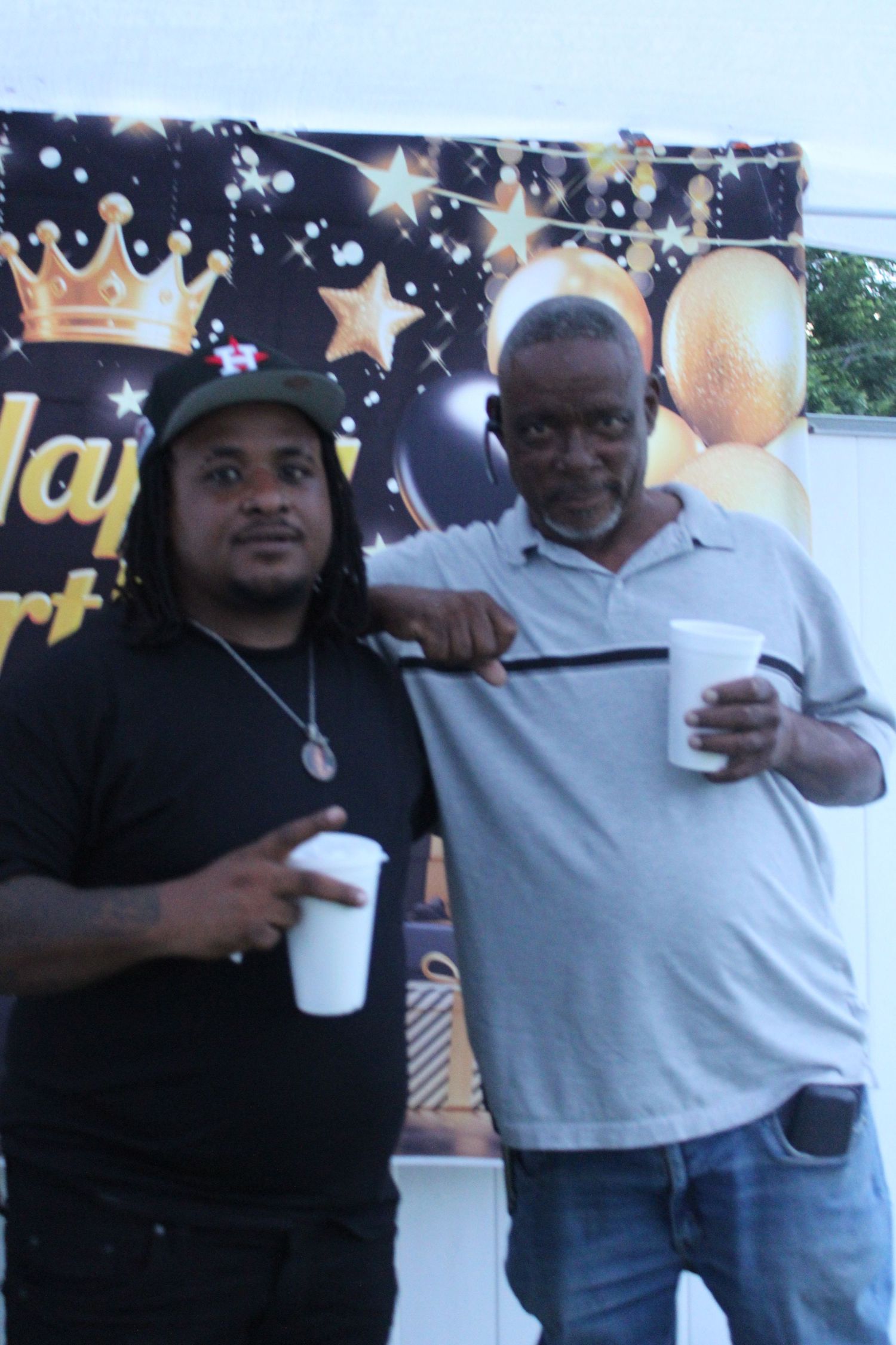 Two men smiling and holding cups in front of a Happy Birthday banner at a party.