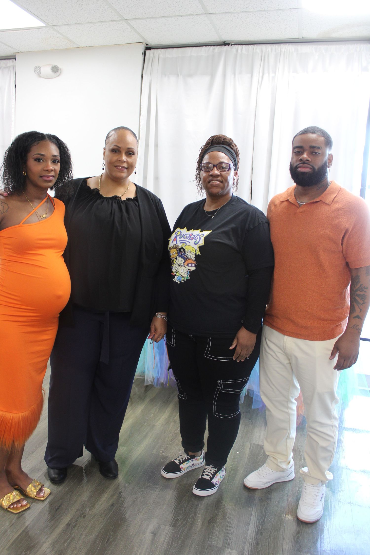 Four people pose together at baby shower event, pregnant woman in orange dress on left.