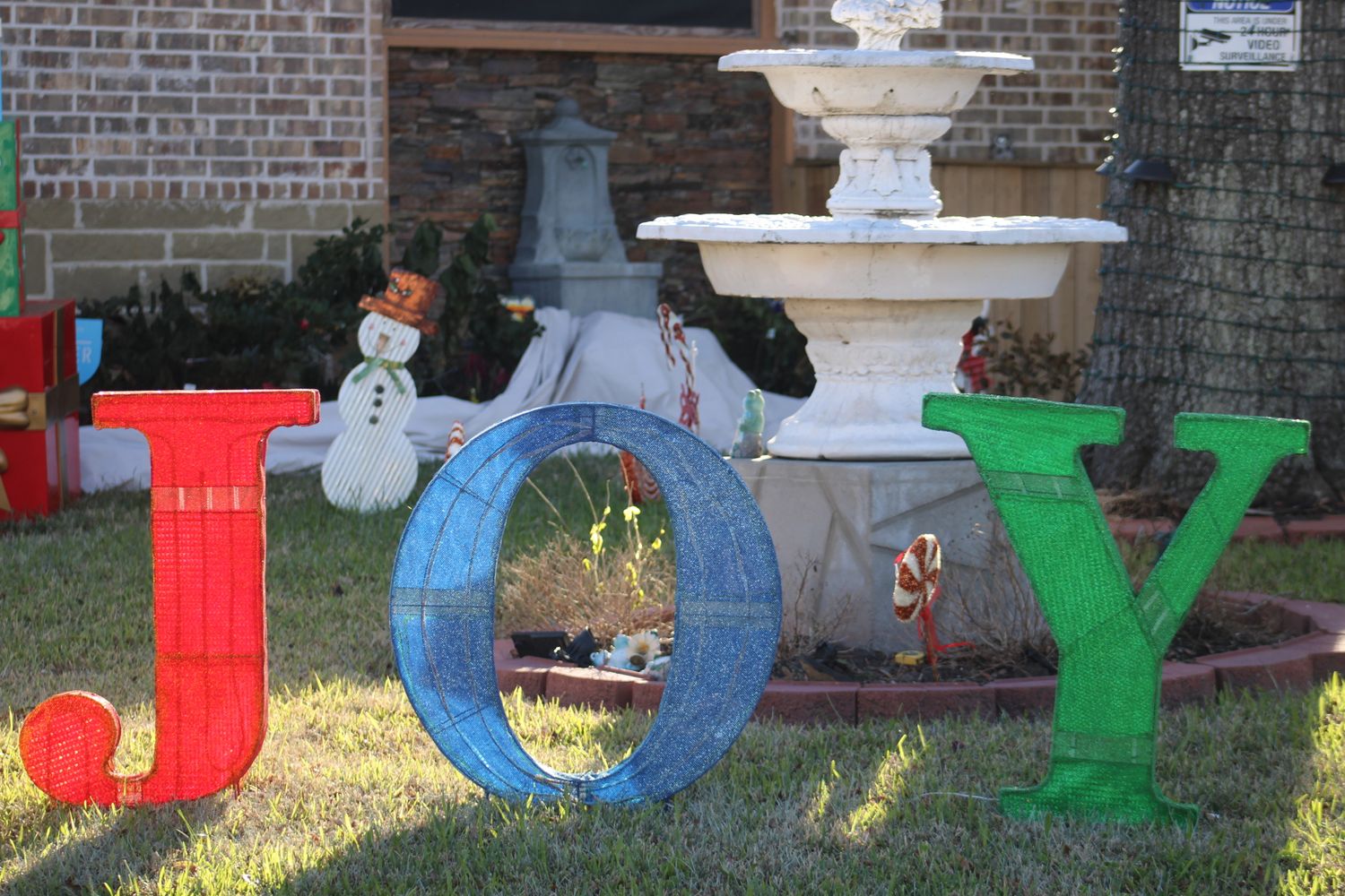 Colorful JOY letters displayed on a front lawn with a snowman and fountain for Christmas.