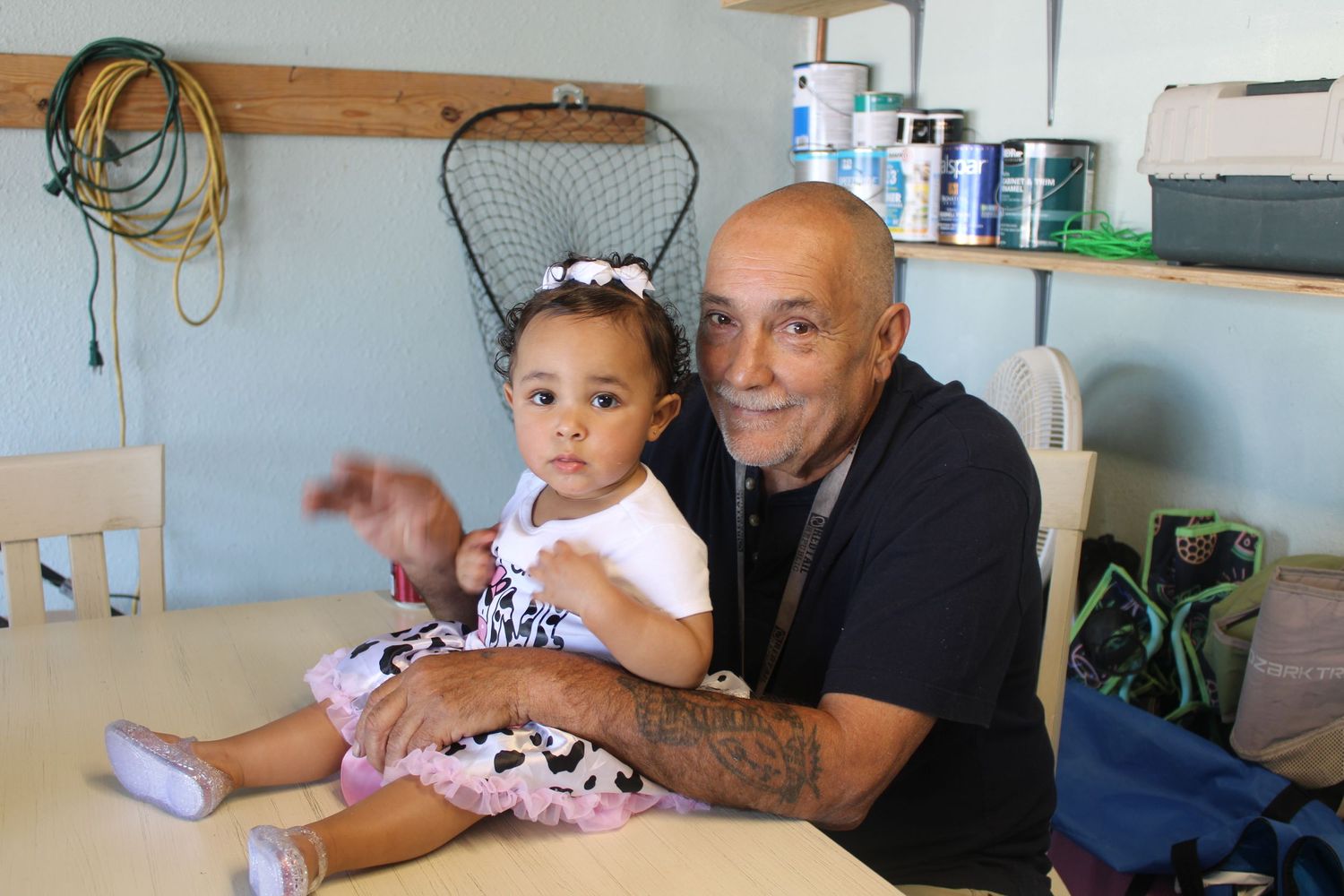 Bald tattooed grandfather smiling while holding baby girl in pink outfit at indoor table.