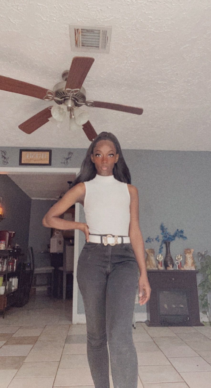 Slim woman in white sleeveless top and gray jeans poses indoors beneath ceiling fan.