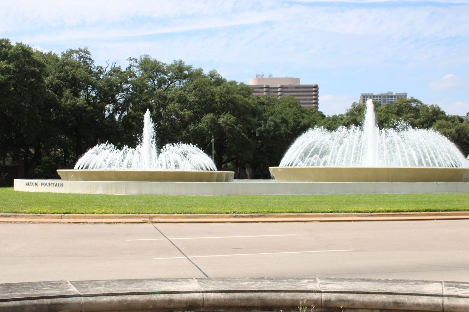 Large decorative water fountains spraying water in a green park with city buildings visible in background.